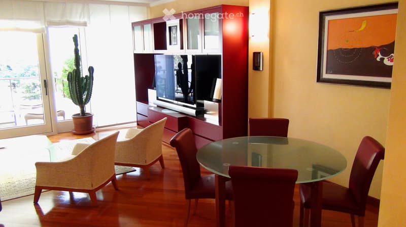 Dining area with a glass table, red chairs, white armchairs, a cactus plant, and a TV cabinet with a framed artwork on the wall.