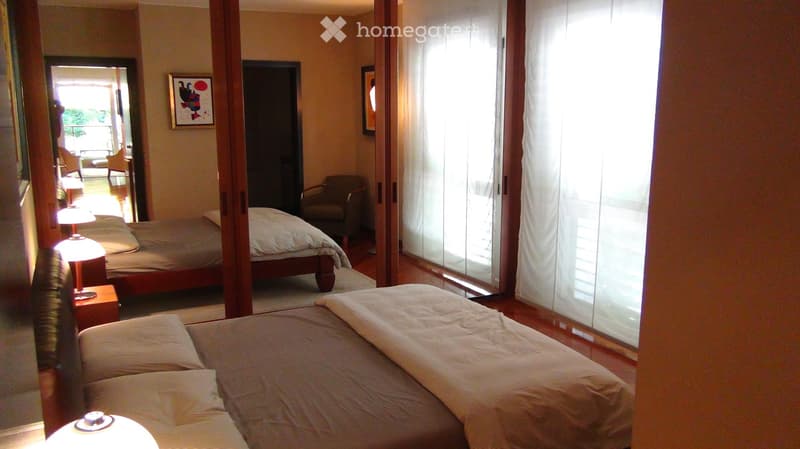 Spacious bedroom with three beds, nightstands, lamps, a chair, and a large mirror. The room features wooden flooring and large windows with sheer curtains.