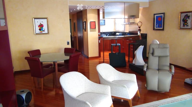 Open-plan living and dining area with wooden flooring, glass dining table with red chairs, white armchairs, a recliner chair, and a kitchen island with stools. Artwork adorns the walls, and a suitcase is placed near the kitchen.