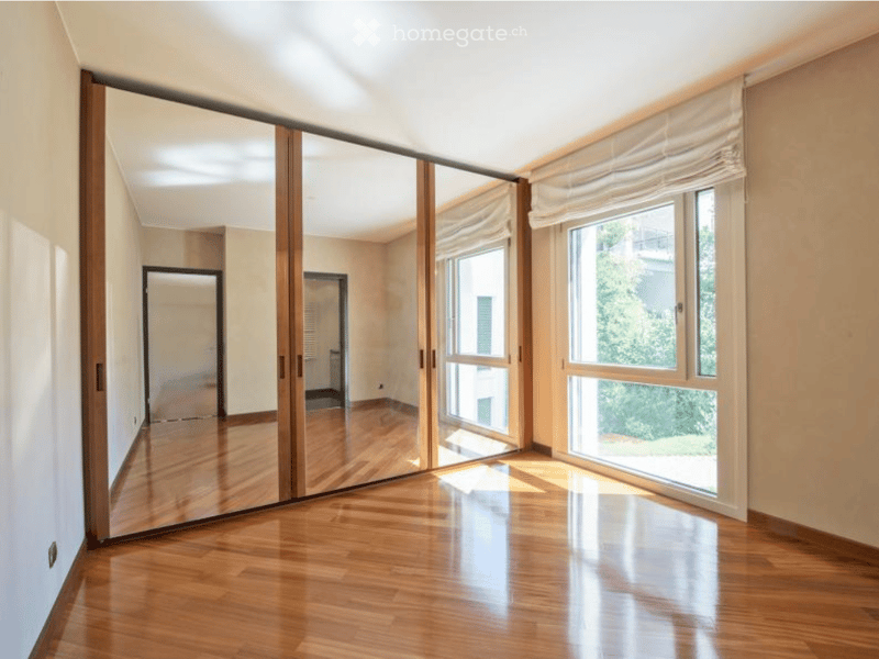 Empty hallway with wooden flooring, large mirrored sliding doors, and large windows with white blinds.