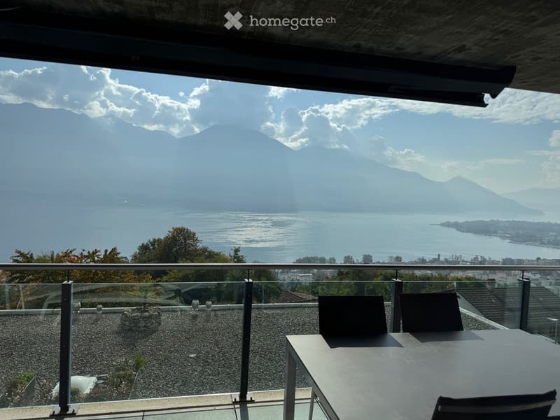 Roof terrace with a panoramic view of a lake and mountains, featuring a glass railing, a table, and chairs.