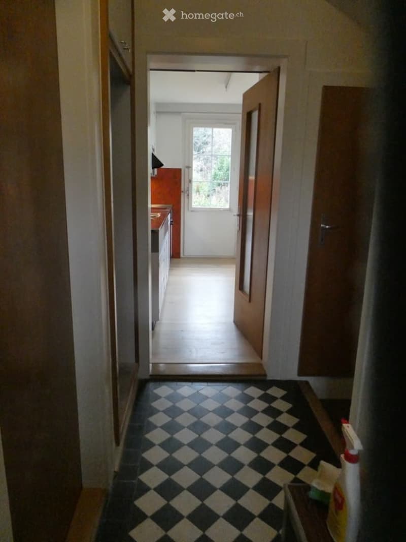 A narrow hallway with checkered black and white tiles, leading to a kitchen area with white cabinets and a window. The hallway has wooden doors on either side and a partially open door at the end.