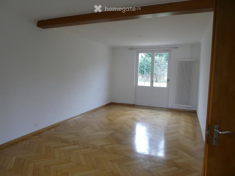 Empty room with wooden herringbone flooring, white walls, large window with white frame, radiator, and wooden door frame.