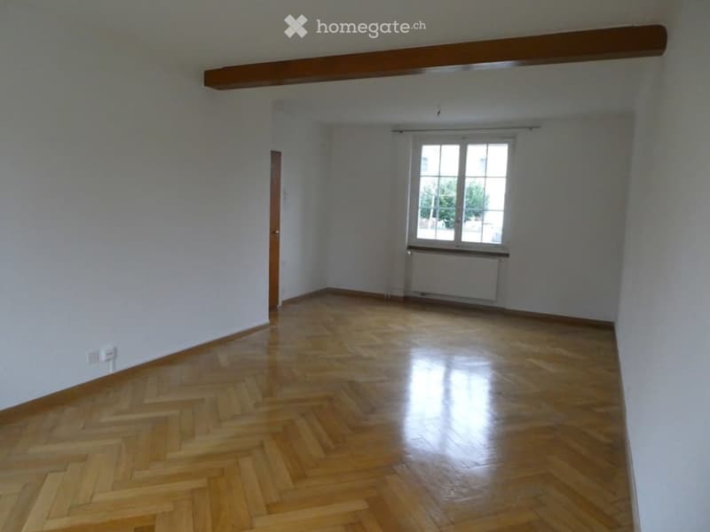 Empty room with wooden herringbone flooring, white walls, and a large window with white frame and radiator below. Exposed wooden beam on ceiling.