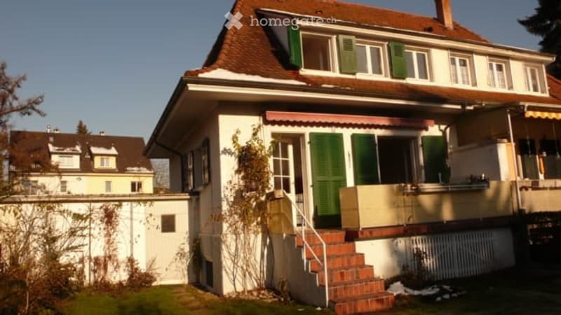 Two-story house with a red-tiled roof, white walls, green shutters, and a small balcony accessible from the upper floor.