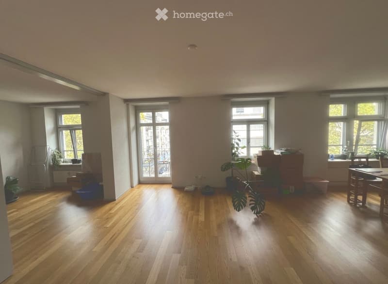 Bright, unfurnished room with wooden flooring, large windows, and a balcony door leading outside. A small table and chairs are positioned near the windows, along with several potted plants.