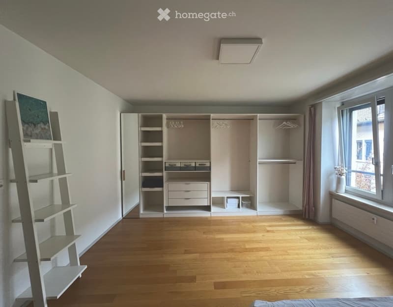 Empty bedroom with wooden flooring, built-in wardrobe with shelves and drawers, a ladder shelf, and a window with a vase on the sill.