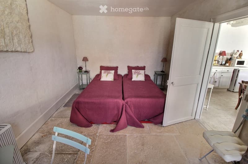 Cozy bedroom with two single beds, red bedding, nightstands, lamps, and a view into an adjacent kitchen area.