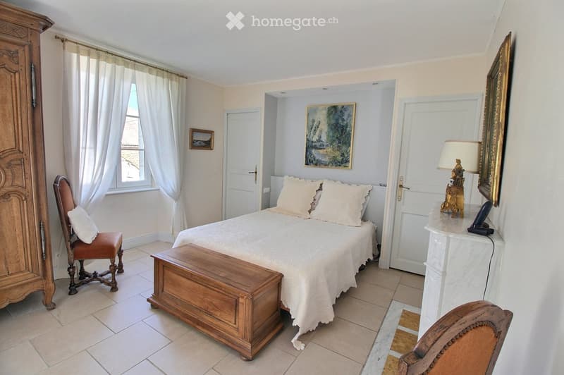 Elegant bedroom with a large bed, wooden chest, antique chairs, a nightstand with a lamp, and framed artwork on the walls.