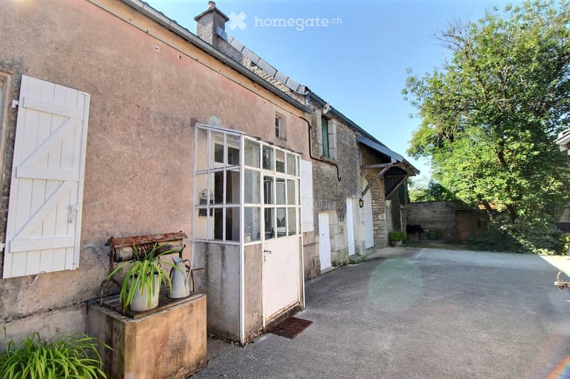 A rustic stone building with a white shuttered window, a large glass-paneled door, and a small bench with a potted plant and a metal pitcher. The building has a chimney and is surrounded by greenery and a gravel driveway.