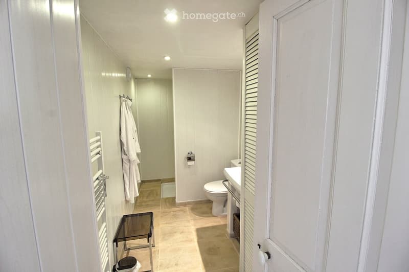 Modern bathroom with white tiles, a toilet, sink, towel rack, and a bathrobe hanging on the wall.