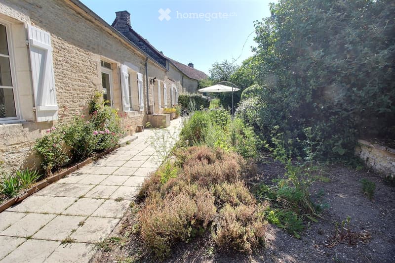 Garden path leading to a stone house with white shutters, surrounded by greenery and flowers.