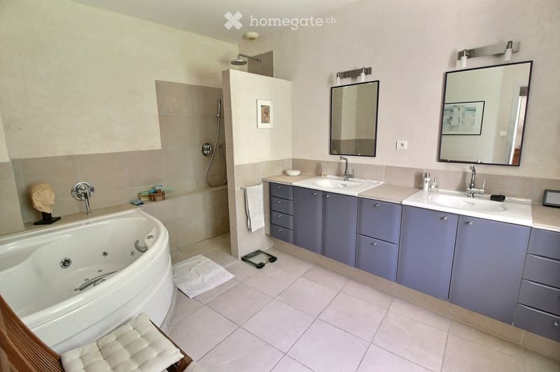 Modern bathroom with a large bathtub, double sink vanity with grey cabinets, mirrors, and a shower area with a glass enclosure.