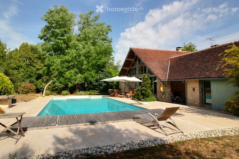 Outdoor area with a swimming pool, wooden deck, lounge chairs, umbrella, and dining set under trees.