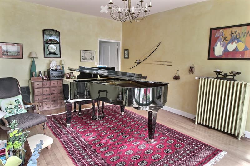 A well-furnished room with a grand piano, a red patterned rug, a wooden chair, a small table, a lamp, and a radiator. The walls are painted in a light yellow color, and there are several picture frames and decorative items on display.
