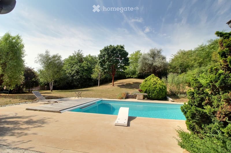 A well-maintained garden with a swimming pool, surrounded by trees and shrubs, featuring lounge chairs and a small table on the deck.
