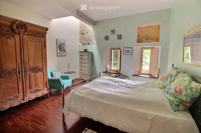 Cozy bedroom with wooden floors, a large bed with a white duvet and colorful pillows, a teal armchair, a small table with a lamp, a wooden wardrobe, and a drawer unit. The room features exposed stone walls and large windows with shutters.