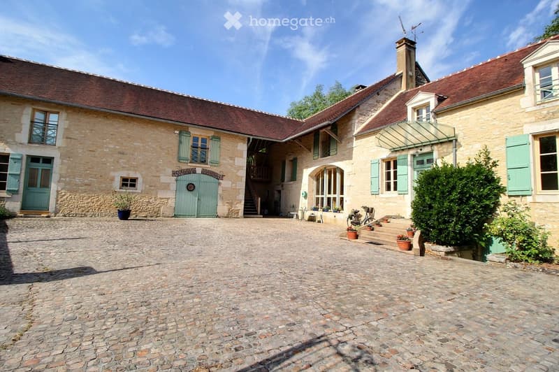 A two-story stone building with a red-tiled roof, green shutters, and a central courtyard with a cobblestone floor. The building features multiple windows, a staircase leading to an upper level, and several potted plants near the entrance.