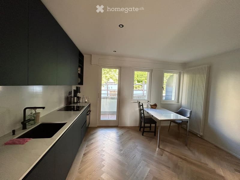 Modern kitchen with light wooden flooring, dark grey cabinets, a sink with a black faucet, a coffee maker, and a small dining area with a table and chairs.