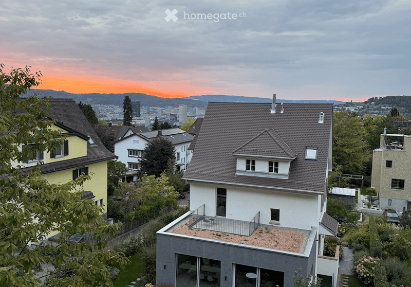 Modern multi-story house with a flat roof terrace, surrounded by greenery and other residential buildings, with a scenic cityscape view in the background.