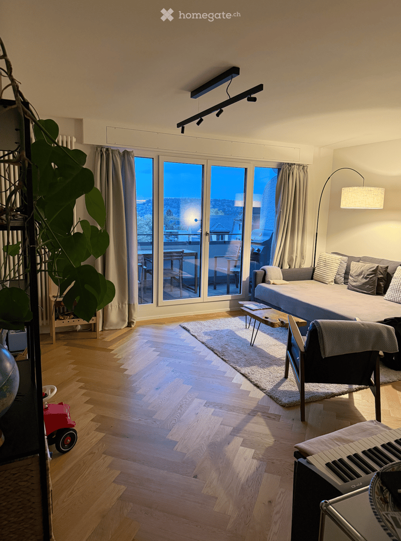 Modern living room with wooden flooring, large windows offering a view, a sofa, coffee table, chairs, and a small table on the balcony.