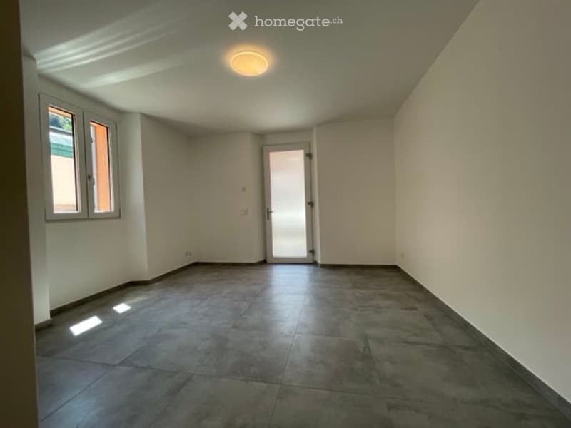 Empty room with tiled floor, white walls, and a single door leading to another space. Natural light streams in from a window on the left side.