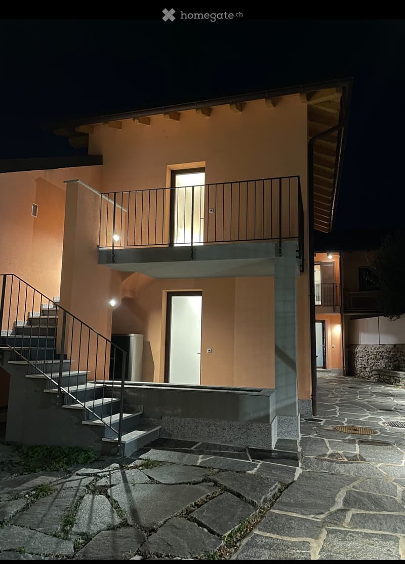 Two-story building with a raised ground floor, stone-paved pathway, and illuminated entrance.