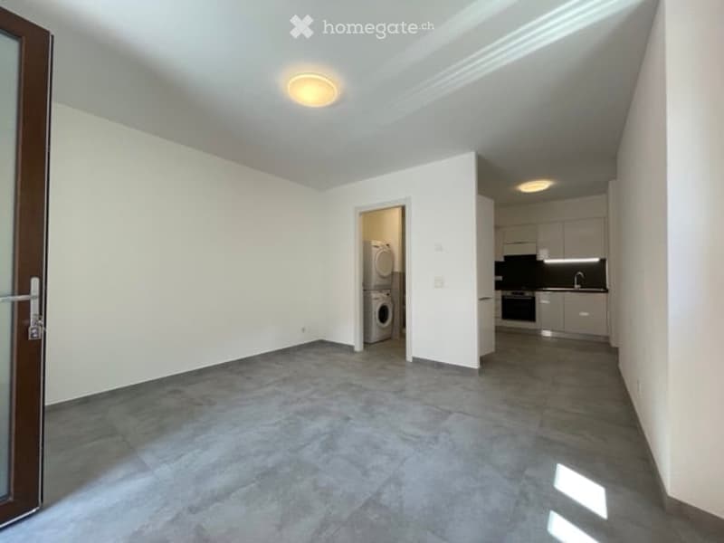Empty room with grey tiled floor, white walls, and a doorway leading to a kitchen area with cabinets and appliances.