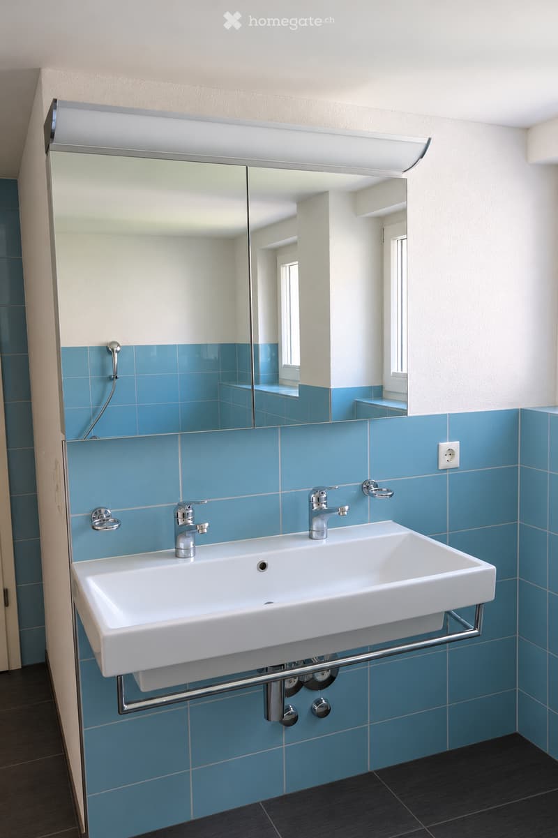 Modern bathroom with a white sink, dual faucets, a large mirror with built-in lighting, and blue tiled walls.
