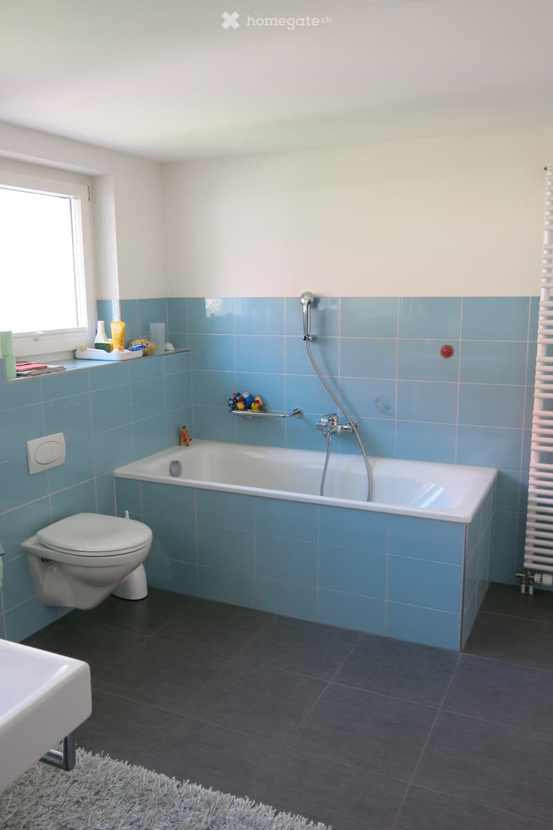 Modern bathroom with blue tiles, bathtub, toilet, sink, and a window with toiletries on the sill.