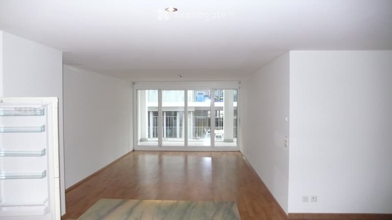 Empty room with wooden flooring, white walls, large sliding glass doors leading to a balcony, and a small rug on the floor.