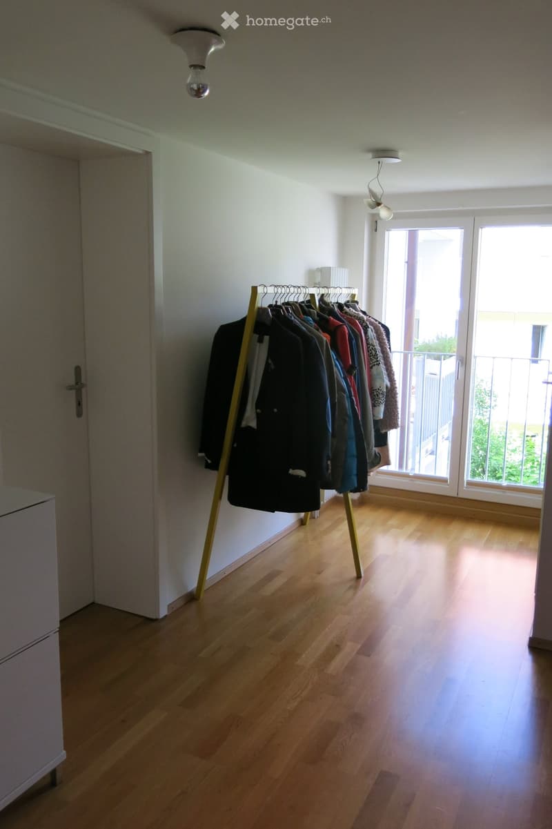 Empty room with light wooden flooring, white walls, and large windows with a view of greenery. A coat rack with several jackets is positioned against the wall.