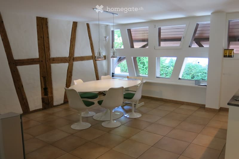 Bright dining area with a round white table surrounded by white chairs with green cushions, pendant lights hanging above, and large windows offering natural light.