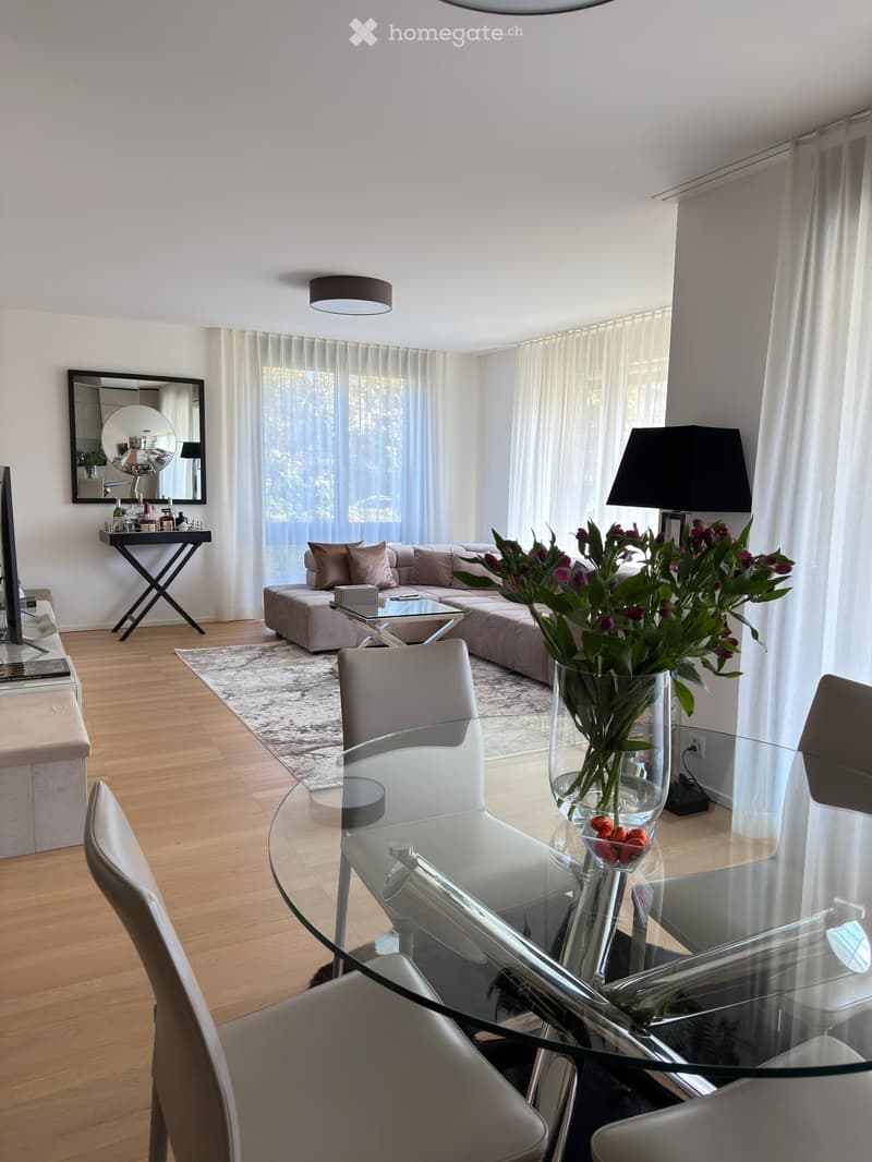 Modern living and dining area with light wood flooring, a grey sofa, a glass coffee table, a glass dining table with chairs, a vase with flowers, a mirror, and a lamp.