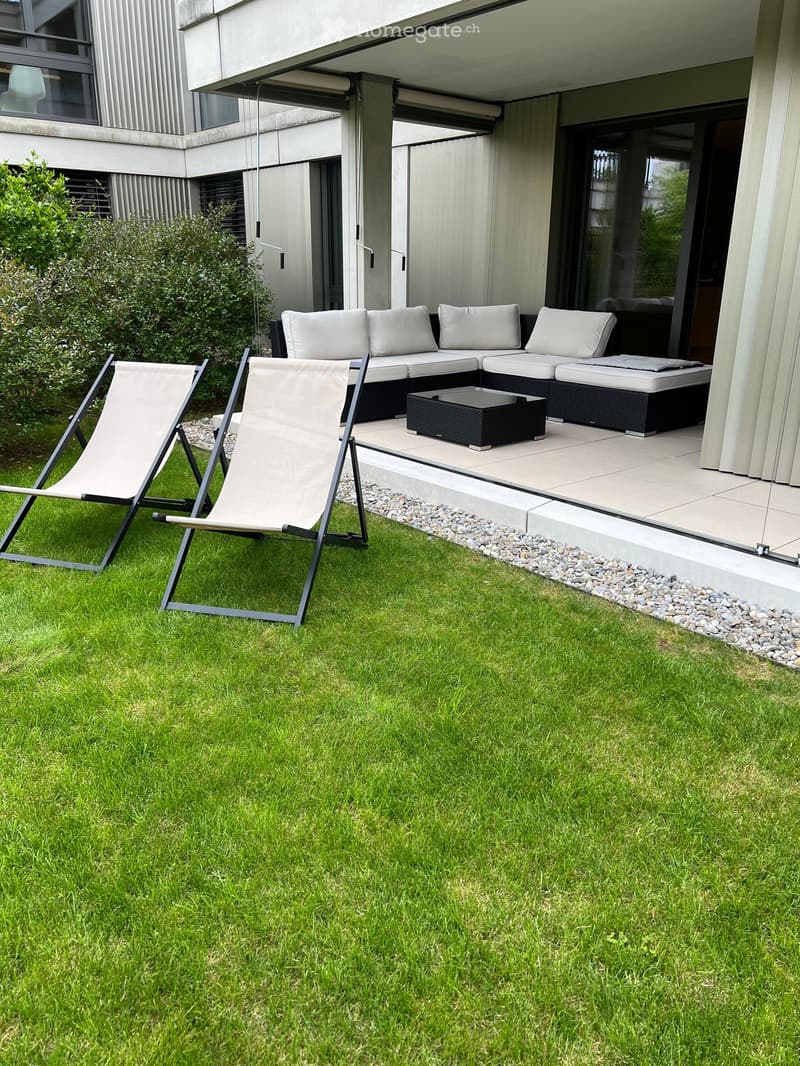 Well-maintained lawn with two lounge chairs and a modern outdoor sofa set with a coffee table on a tiled patio area.