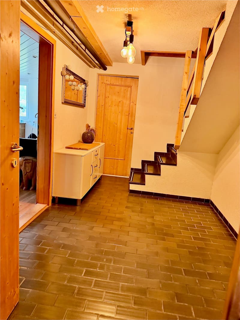 A narrow hallway with wooden flooring, a small cabinet with drawers, a decorative mirror, and a vase on top of the cabinet. The hallway leads to a room with visible furniture.