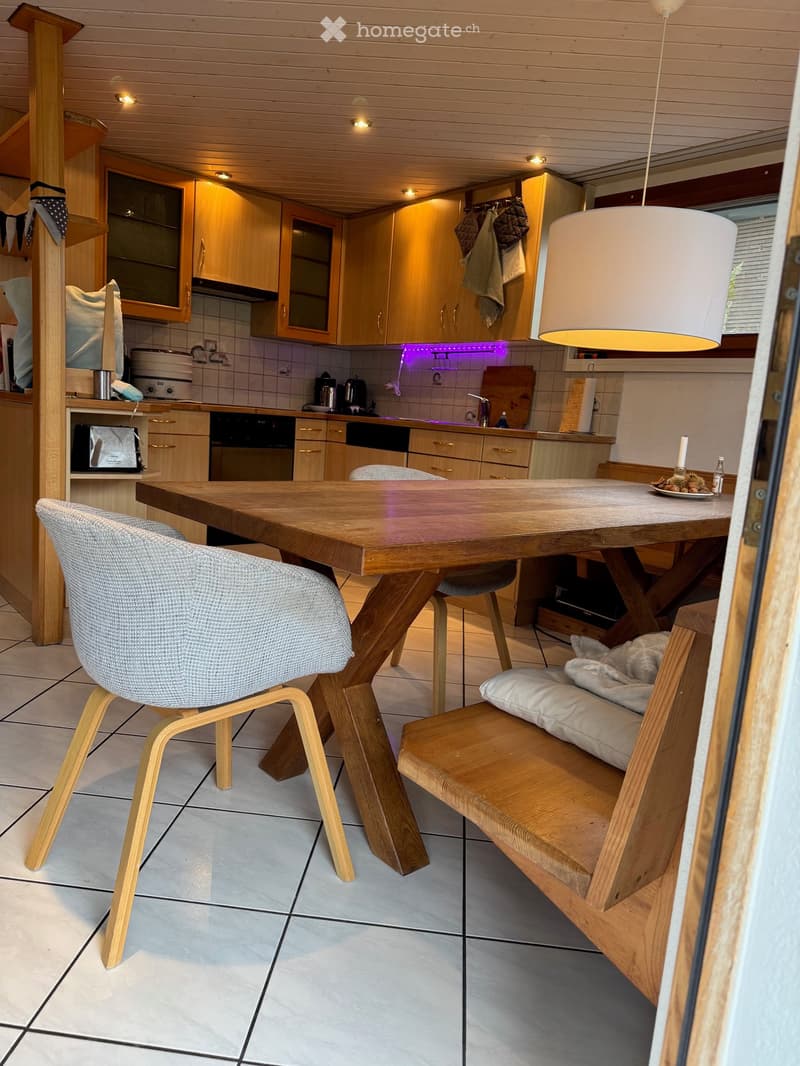 Modern kitchen with wooden cabinets, a large wooden dining table, a bench, and a chair. The kitchen features a hanging lamp, a tiled floor, and various appliances including an oven and a fridge.