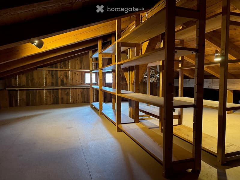 An empty attic space with wooden beams, exposed rafters, and built-in wooden shelves. The room has a concrete floor and is illuminated by warm lighting from spotlights.