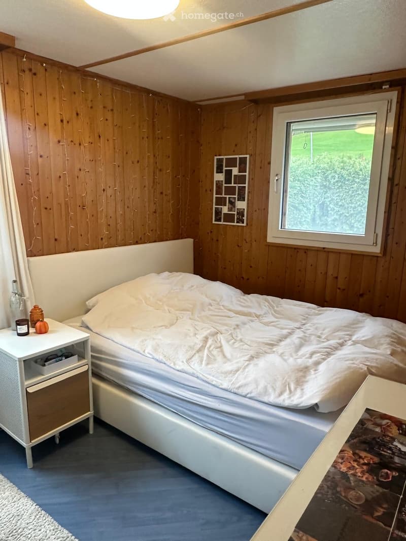 Cozy bedroom with wooden walls, a large bed with white bedding, a nightstand with candles and a bottle, and a window with a view of greenery.