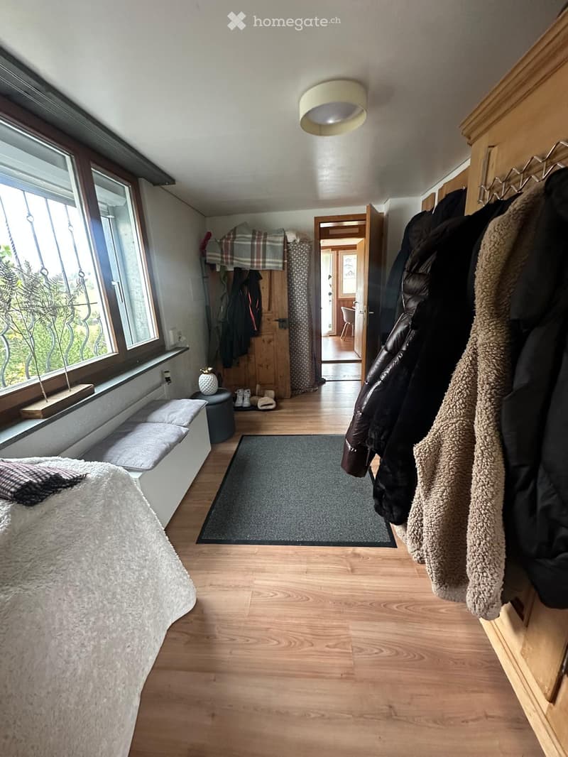 A cozy, well-lit room with wooden flooring, a large window, a bed with a fluffy white duvet, a bench seat, a coat rack with jackets, a small rug, and a stool with shoes.