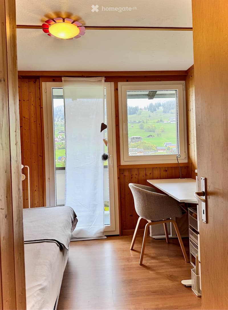 Cozy room with wooden walls and floor, featuring a bed, desk, chair, and a large window overlooking a scenic landscape.