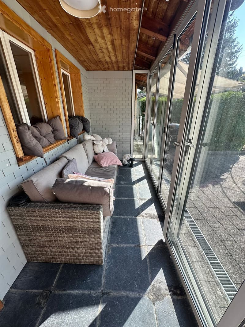 Balcony with a grey sofa, wicker basket, and decorative pillows, featuring large glass doors and a wooden ceiling.