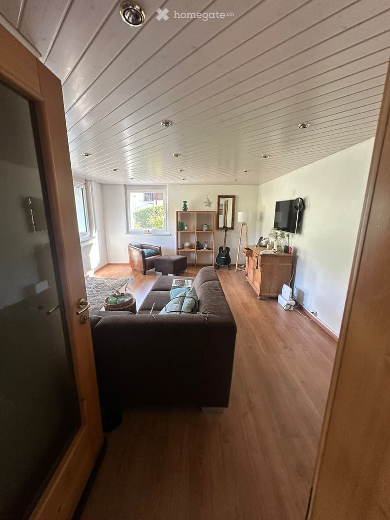 Living room with wooden flooring, slanted ceiling, large windows, sofa, armchair, ottoman, small table, guitar, shelf, mirror, and television.