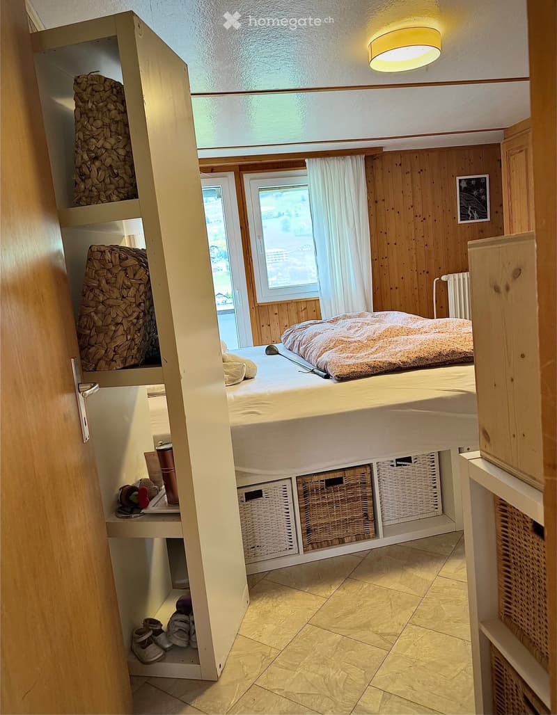 Cozy bedroom with wooden walls, a bed with a patterned blanket, storage baskets under the bed, a small table with a cup, and a radiator near the window.