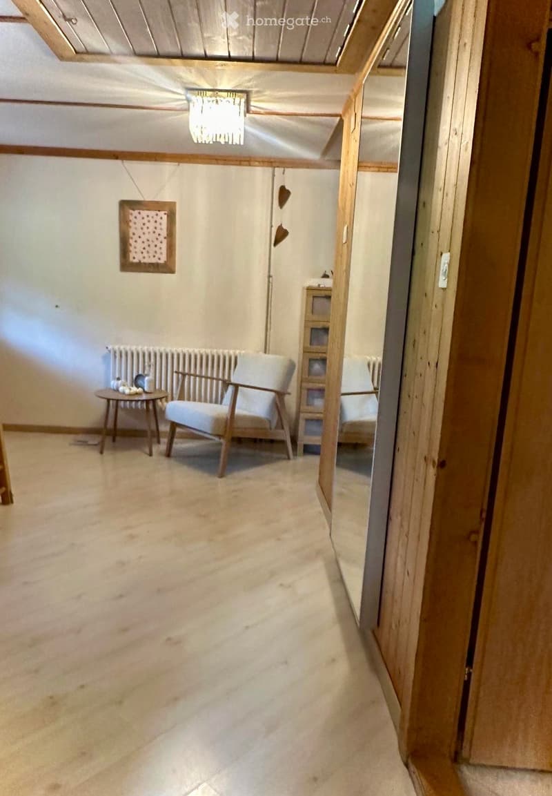A furnished room with a light-colored carpet, a white armchair, a small round table, a radiator, and a wooden cabinet. The room has exposed wooden beams on the ceiling and a partially visible adjacent room.