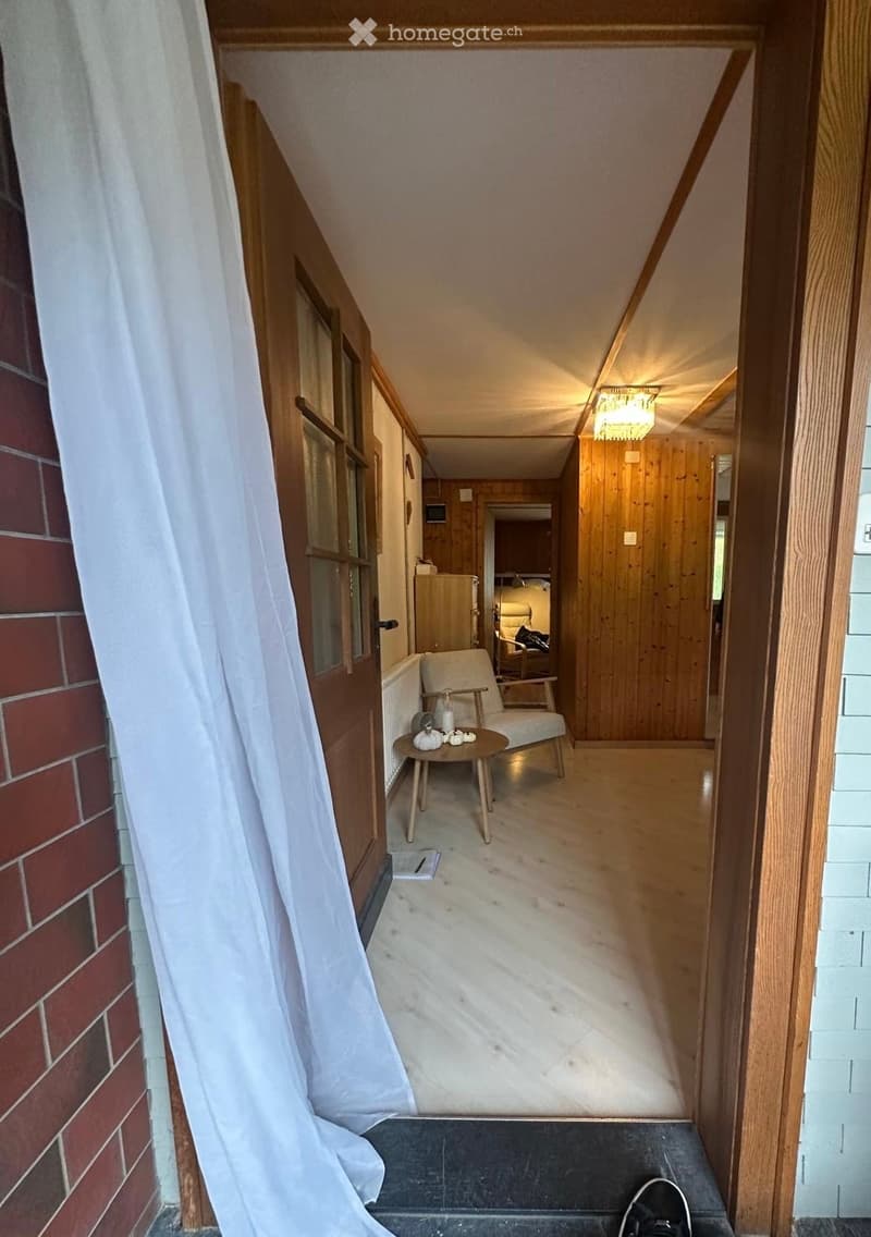 A narrow hallway with wooden walls and flooring, featuring a small round table with a chair, a chandelier, and a glimpse of a bedroom area with a bed and wardrobe.