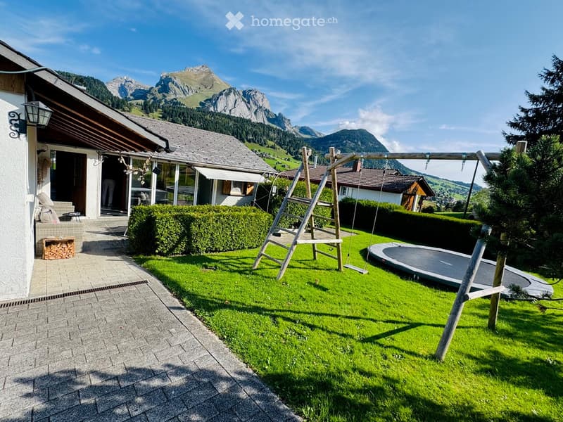 Lush green garden with a swing set, trampoline, neatly trimmed hedges, and a view of snow-capped mountains in the background.