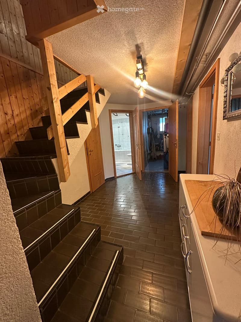 A well-lit hallway with wooden stairs, tiled floor, and open doors leading to other rooms, including a bathroom and a living area.