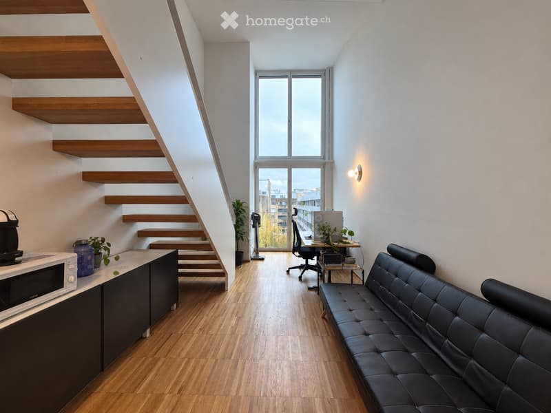 Modern, open-plan living and dining area with a black leather sofa, a small kitchen area with a microwave, a wooden staircase leading to an upper level, and a desk with a chair and laptop.