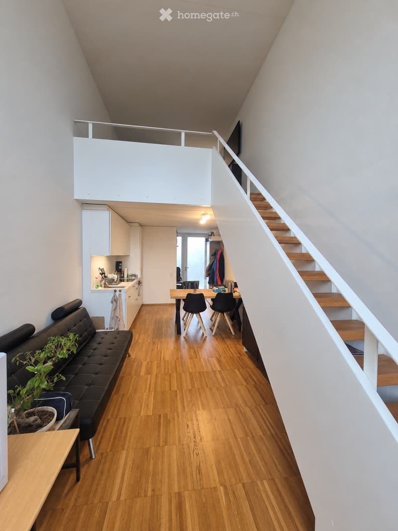 Open-plan living and dining area with wooden flooring, a black leather sofa, a small dining table with chairs, and a modern kitchen area with white cabinets and appliances.